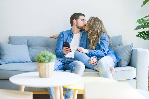 Jeune couple amoureux étreignant assis sur le canapé en utilisant smartphon
 - Photo, image