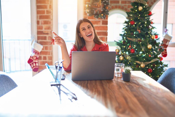 Beautiful woman sitting at the table working with laptop at home around christmas tree with a big smile on face, pointing with hand finger to the side looking at the camera. - 写真・画像