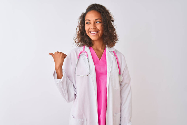Joven doctora brasileña con estetoscopio de pie sobre un fondo blanco aislado sonriendo con la cara feliz mirando y señalando hacia un lado con el pulgar hacia arriba
. - Foto, imagen