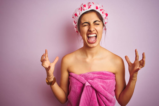 junge schöne Frau trägt ein Duschtuch nach dem Bad über rosa isolierten Hintergrund verrückt und verrückt schreien und schreien mit aggressivem Ausdruck und erhobenen Armen. Frustrationskonzept. - Foto, Bild
