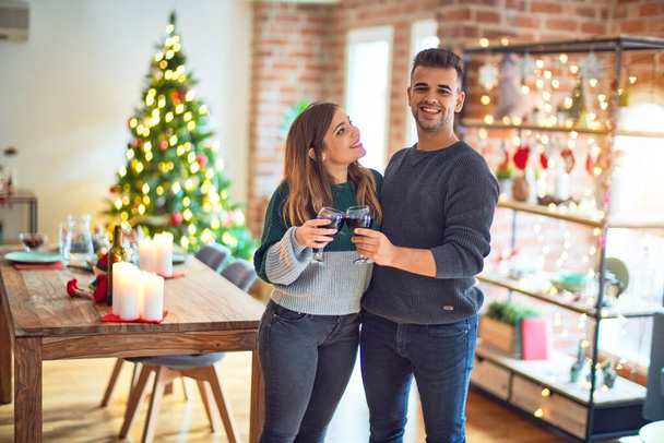 Νεαρό όμορφο ζευγάρι χαμογελά ευτυχισμένη και αυτοπεποίθηση. Πρόποση με ένα φλιτζάνι κρασί που γιορτάζει τα Χριστούγεννα στο σπίτι - Φωτογραφία, εικόνα