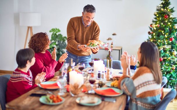 Krásná rodina se usmívá šťastně a sebevědomě. Jeden z nich stojí a ukazuje pečeného krocana, jak doma slaví Vánoce. - Fotografie, Obrázek