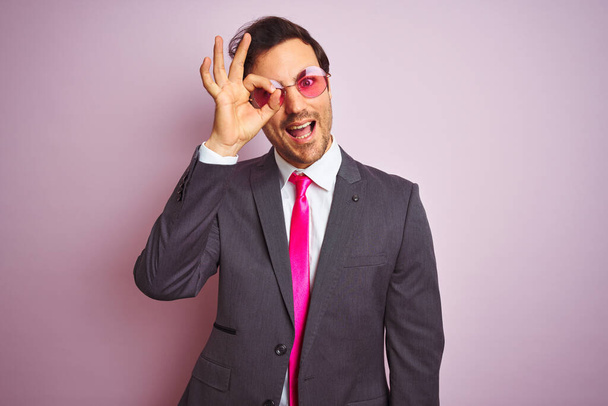 Молодой красивый бизнесмен в костюме и солнцезащитных очках на изолированном розовом фоне делает хорошо жест с улыбкой на руке, глаза смотрят сквозь пальцы с счастливым лицом
. - Фото, изображение