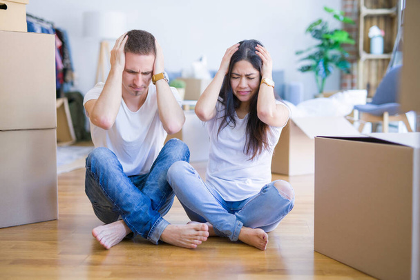 Young beautiful couple sitting on the floor at new home around cardboard boxes suffering from headache desperate and stressed because pain and migraine. Hands on head. - Photo, Image
