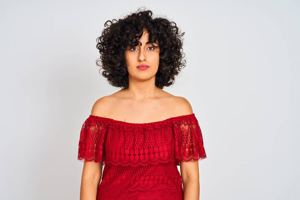 Young arab woman with curly hair wearing red dress standing over isolated white background with serious expression on face. Simple and natural looking at the camera. - Фото, зображення