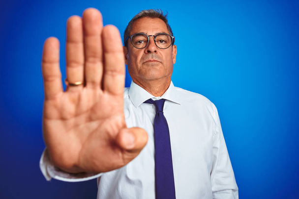 Красивый бизнесмен среднего возраста в очках, стоящих на изолированном синем фоне с открытой рукой делает знак стоп с серьезным и уверенным выражением лица, защитный жест
 - Фото, изображение