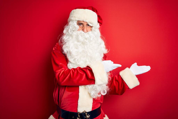 gutaussehender Mann mittleren Alters im Weihnachtsmannkostüm, der vor isoliertem rotem Hintergrund steht und einlädt, lächelnd und natürlich mit offener Hand einzutreten - Foto, Bild