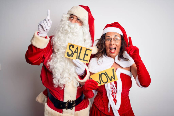 Pari yllään Santa puku tilalla wow ja myynti banneri yli eristetty valkoinen tausta hymyillen hämmästynyt ja yllättynyt ja osoittaa ylös sormet ja nosti kädet
. - Valokuva, kuva