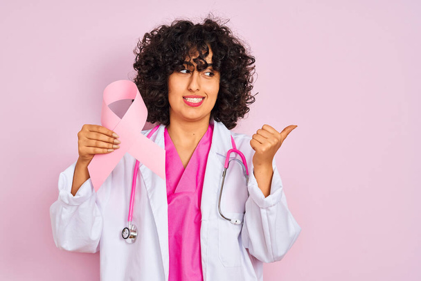 Young arab doctor woman with curly hair holding cancer ribbon over isolated pink background pointing and showing with thumb up to the side with happy face smiling - 写真・画像