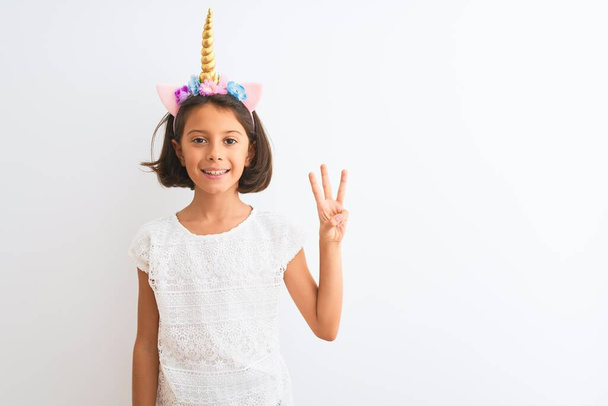mooi kind meisje dragen eenhoorn diadeem staande over geïsoleerde witte achtergrond tonen en wijzen met vingers nummer drie terwijl glimlachen zelfverzekerd en gelukkig. - Foto, afbeelding