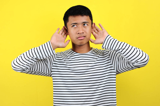 Mladý pohledný Asiat v pruhovaných tričkách poslouchá něco s rukou přes ucho a poslouchá drby nebo drby. Koncept hluchoty - Fotografie, Obrázek