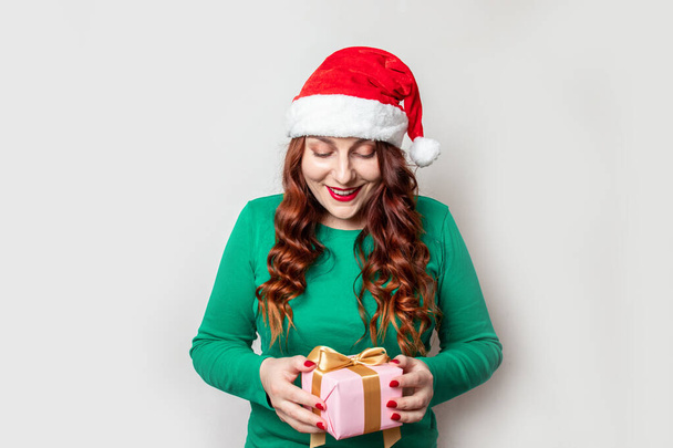 Щаслива молода кучерява дівчина з капелюхом Санти, зелений светр тримає сувенірну коробку з золотим луком, яка виглядає здивованою на сірому тлі. Веселого Різдва і Веселого Нового Року. Різдво. - Фото, зображення