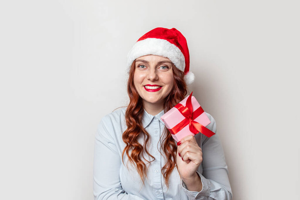 Santa Claus chica con el pelo rizado y un sombrero rojo con un bumbon sostiene una caja de regalo con un lazo de cinta de satén rojo y sonríe sobre un fondo de pared gris. Feliz Navidad y Año Nuevo banner web para el sitio
. - Foto, imagen