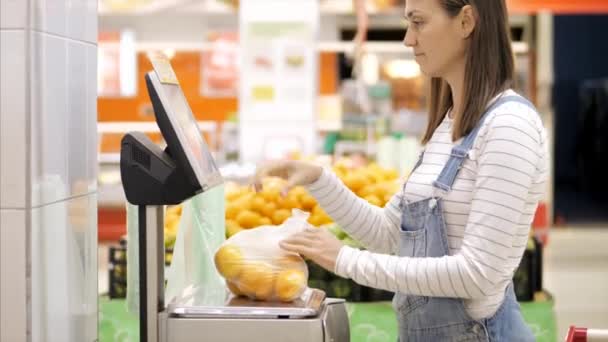 Young woman in denim overall is weighs fresh oranges at grocery store. - Felvétel, videó