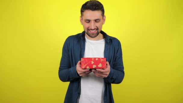 A young man in a white T-shirt admires the box with a gift in his hands, smiles and looks at the camera, yellow background. Holidays concept: New Year, Christmas, Valentine's Day birthday. 4k footage - Felvétel, videó
