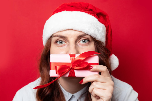 Pretty curly red-haired lady in santa hat expressing happy emotions and holding present box with satin ribbon bow on red background - Zdjęcie, obraz