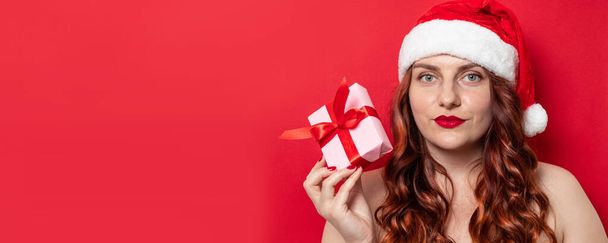 schöne Frau mit Weihnachtsmannhut hält Geschenk-Box mit Satinband Schleife Blick in die Kamera auf rotem Hintergrund - Foto, Bild