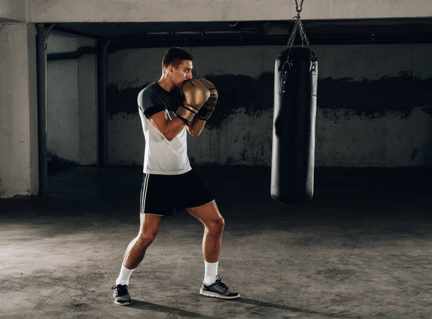 Muscular young man in boxing gloves and shorts shows the different movements and strikes on a dark background - Photo, Image