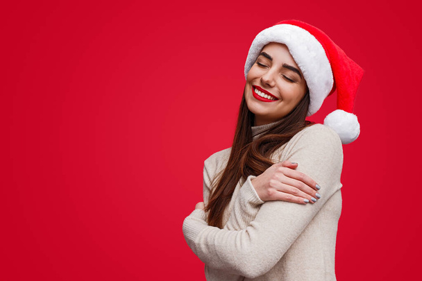 Charming dreamy woman in Santa hat embracing arms - Foto, afbeelding