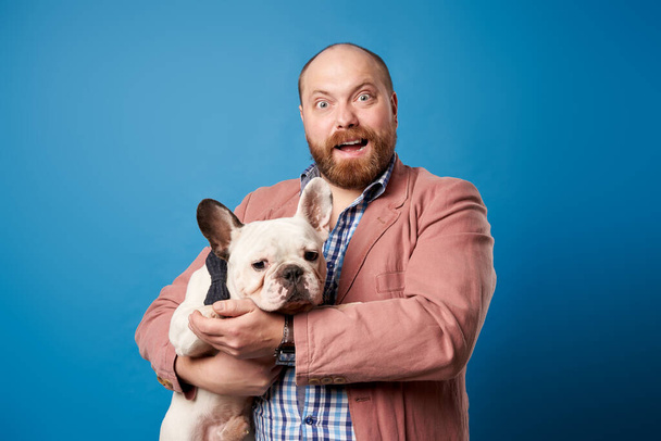 Bearded man with french bulldog in arms on empty blue background. - Foto, imagen