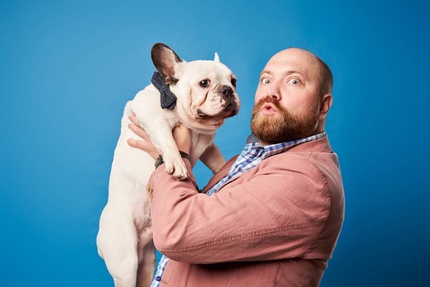 Bald male in jacket with french bulldog in his arms on empty blue background - Photo, Image