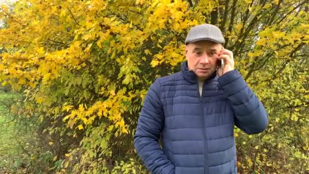 man in a gray cap and a blue jacket in autumn park talking on the phone, gesturing expressively with his hands, the concept of scandalous conversation, quarrels, close-up - Imágenes, Vídeo