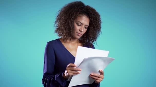 Pretty african american female boss checks documents, utility bills. Serious businesswoman standing on blue background. She is satisfied with work of staff.  - Filmati, video