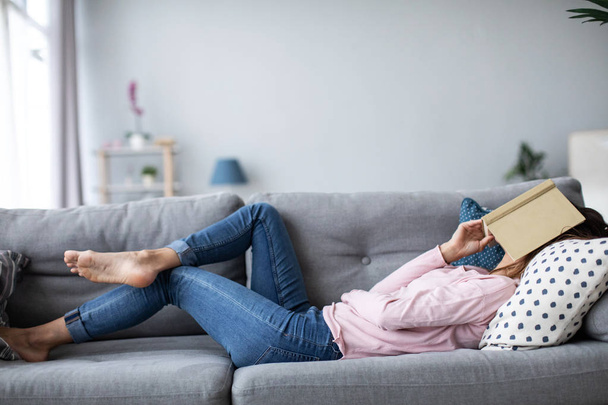 Young beautiful woman with a book while lying on a sofa in the living room.  - Fotó, kép
