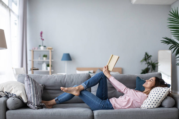 Young beautiful woman is reading a book while lying on a sofa in the living room.  - Fotoğraf, Görsel