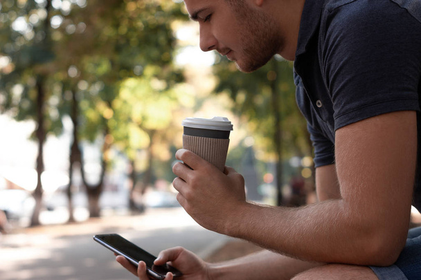 Young man is looking at the screen of his smartphone and drinking coffee in the park. He is wearing a blue t-shirt, blue jeans and white sneakers. Empty space on the screen for your design. Horizontal - Foto, imagen