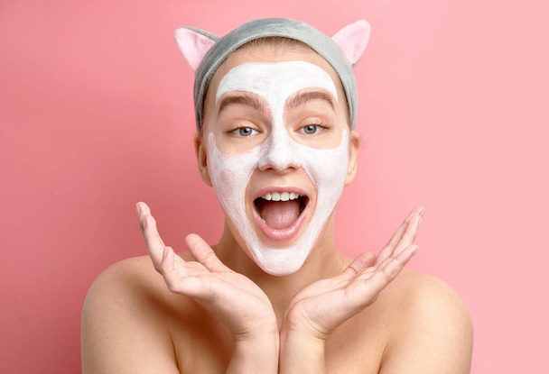 Optimist shocked girl with white cosmetic cleansing face mask and Korean cat ears headband raises palms, smiles joyfully, surprised on pink background - Zdjęcie, obraz