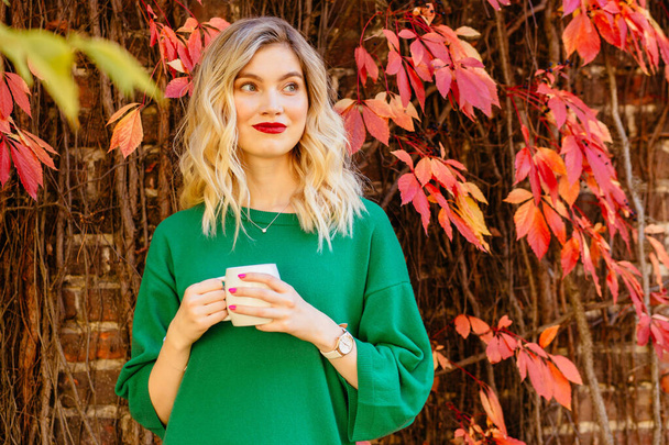 Close-up portret van jonge blonde vrouw met rode lippen drinkt koffie in de herfst buiten, het dragen van warm groene trui, gelukkige levensstijl, herfst concept. Vrouwelijke dreamimg met kopje thee op rode achtergrond. - Foto, afbeelding