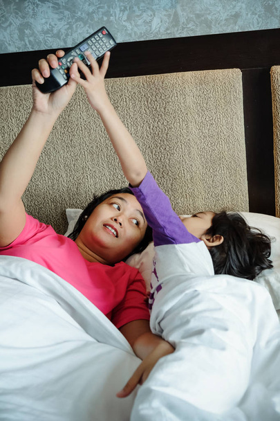 Asiático niño en la cama quiere hacerse cargo de la televisión de control remoto de su madre. Control parental
 - Foto, imagen