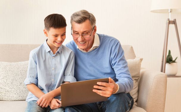 Oğlan ve büyükbaba evdeki koltukta dizüstü bilgisayar kullanıyor. - Fotoğraf, Görsel