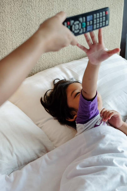Asiático niño en la cama quiere hacerse cargo de la televisión de control remoto de sus manos paternas. Concepto de adicción
 - Foto, imagen