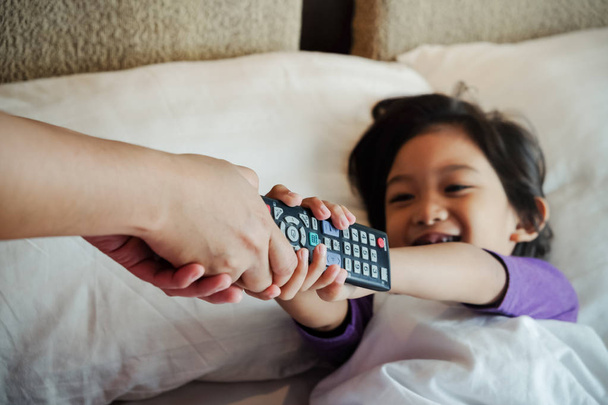 Ázsiai gyermek az ágyon azt akarja, hogy vegye át a televízió távirányító az ő szülői kezét. Függőség vagy szülői tanácsadó koncepció - Fotó, kép