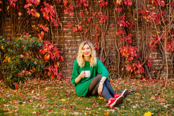 Ρομαντικό όνειρο Ξανθό κορίτσι σε πράσινο πλεκτό πουλόβερ με κούπες στα χέρια στο κόκκινο τοίχο τούβλο με αναρρίχηση σταφύλια στο παρασκήνιο. Φθινοπωρινή Θερμαινόμενη Έννοια. - Φωτογραφία, εικόνα