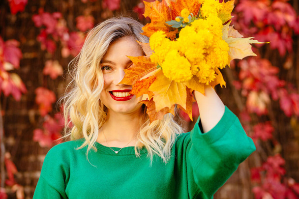 Romantic Dreaming Blond Girl in Green Knitted Sweater with Mug in Hands Sitting on Grass With Colorful Leaves at Red Brick Wall with Climbing Grapes on Background. Autumn Mood Warming Up Concept. - Photo, Image