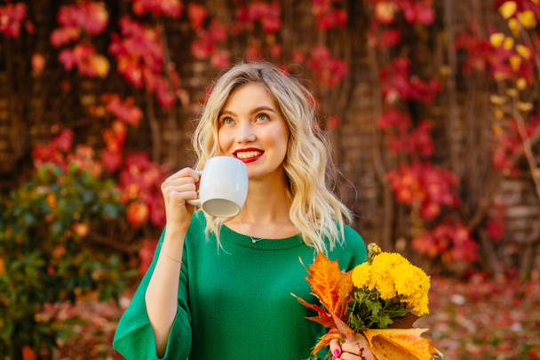 Blond romantic young woman in green sweater holds mug with hot drink and yellow bouguet autumn flowers on red wall with climbing grapes on background. Happy lifestyle, fall concept - Photo, Image