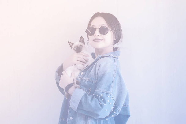 Young attractive woman hugging pussy cat in hands. Cute and glamorous girl in trendy sunglasses posing with her Siamese cat - Фото, зображення