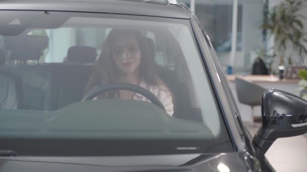 Portret van een jonge, mooie blanke vrouw in de nieuwe auto in de showroom. Zelfverzekerd lachend meisje kiezen auto te kopen. Autohandel, autohandel. - Video