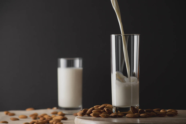 Pouring almond milk into a glass with unfocused glass of milk on background and almond nuts. Almond milk lactose free milk substitute concept. Copy space. - Foto, Imagen