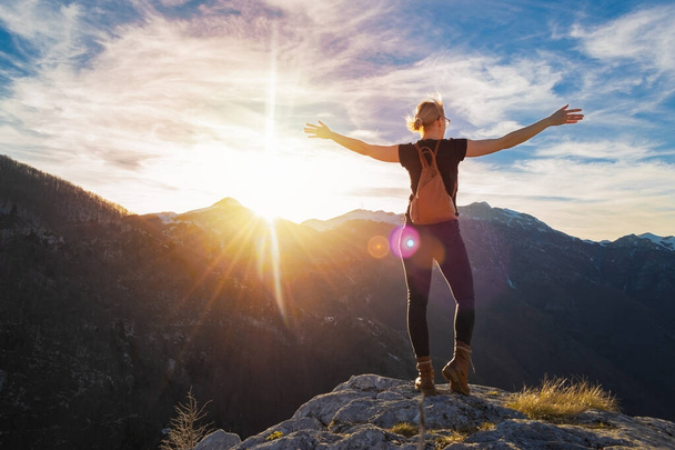Young woman with backpack and spread hands standing on the edge of rocky cliff looking at sunset in the mountains. Motivation and inspiration, freedom concept. Copy space. - Foto, Imagen