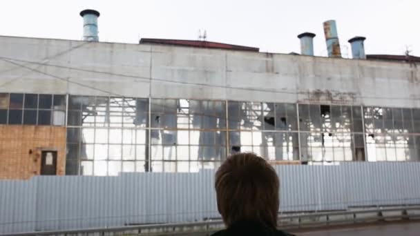 Tylny widok człowieka idącego do ruin opuszczonej starej fabryki. przemysłowy widok na zniszczoną opuszczoną fabrykę do rozbiórki z dużymi rozbitymi szybami, brudnymi zniszczonymi ścianami i zardzewiałymi rurami - Materiał filmowy, wideo