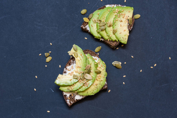 Delicious avocado toast, brown bread, flax seeds, and pumpkin on parchment with a dark background. - Foto, afbeelding