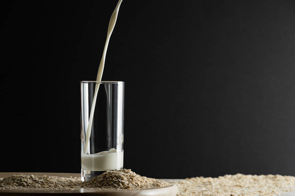 Pouring oat milk into glass on black background and oat flakes on table. Oat milk lactose free milk substitute concept. Copy space, soft shadows. - 写真・画像