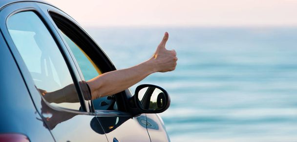 Man showing thumb up from car window with sunset sky and blue sea at background. Copy space on right side. Traveling by car or rent car concept - Photo, image