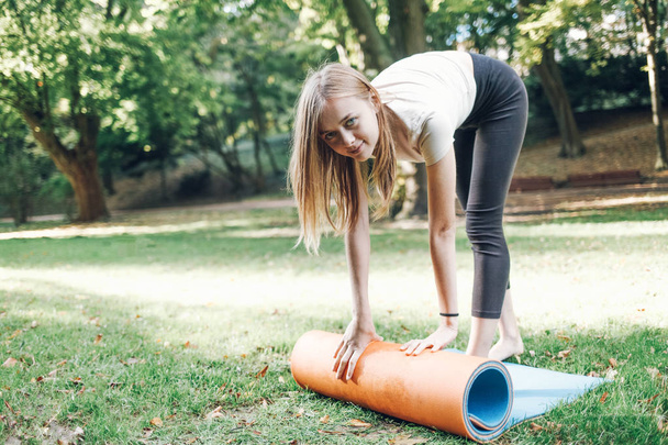 Menina jovem desdobra um tapete de ioga no parque na grama
 - Foto, Imagem