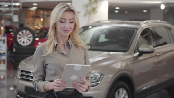 Confident young Caucasian woman using tablet, looking at camera and smiling. Successful blond car dealer posing at the background of new automobiles. Car dealership, car business. - Video