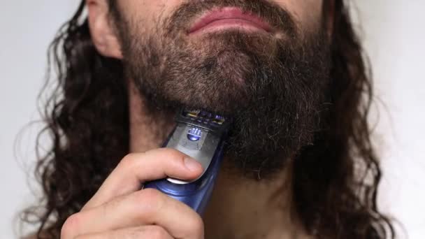 Jóképű, hosszú hajú fiatalember borotválja a szakállát egy nyíróval a fürdőszobában. Közelkép. - Felvétel, videó